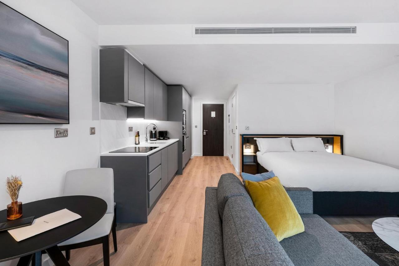 Citysuites 2 Aparthotel Mánchester Exterior foto The photo shows a modern and stylish studio apartment. It features a kitchenette with dark cabinetry and a small dining table with two chairs. There's a cozy sofa with colorful cushions, positioned next to the table. In the background, a large bed wi