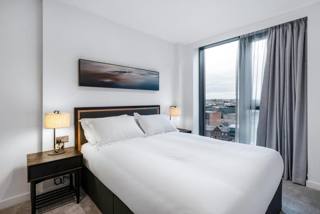 Citysuites 2 Aparthotel Mánchester Exterior foto The image shows a modern bedroom featuring a large bed with a white duvet and two pillows. On either side of the bed are small nightstands with lamps. A large window occupies one wall, draped with light gray curtains, allowing natural light to enter 