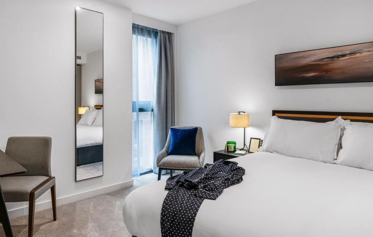 Citysuites 2 Aparthotel Mánchester Exterior foto The photo shows a modern hotel room. It features a neatly made bed with white linens and a dark-colored throw draped over it. There is a small bedside table with a lamp next to the bed. A comfortable chair in a light color is positioned near the wind