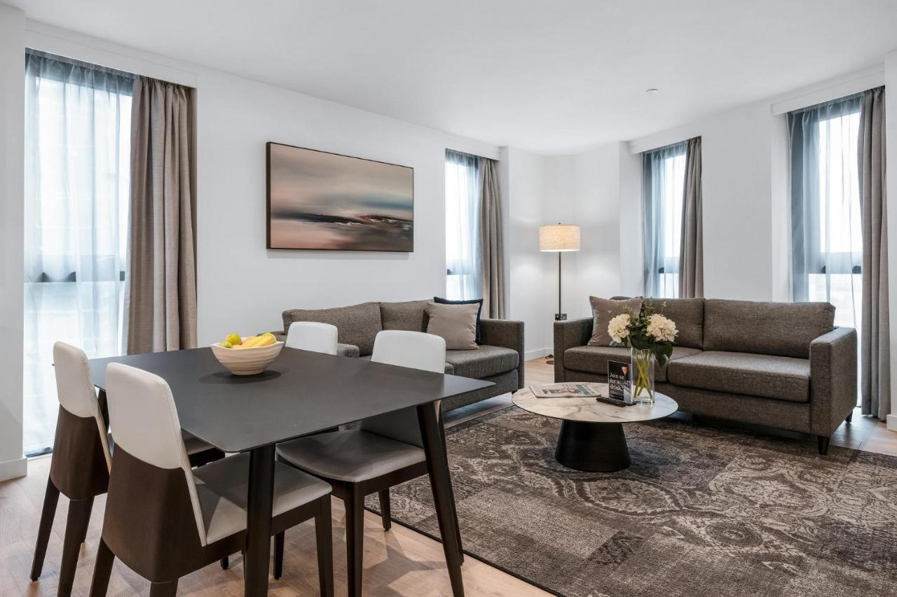 Citysuites 2 Aparthotel Mánchester Exterior foto The photo depicts a modern and stylish interior of a living space. On the left, there is a dining table with four chairs, featuring a bowl of fruits on the table. In the background, there's a cozy seating area with a gray sofa and a smaller round cof
