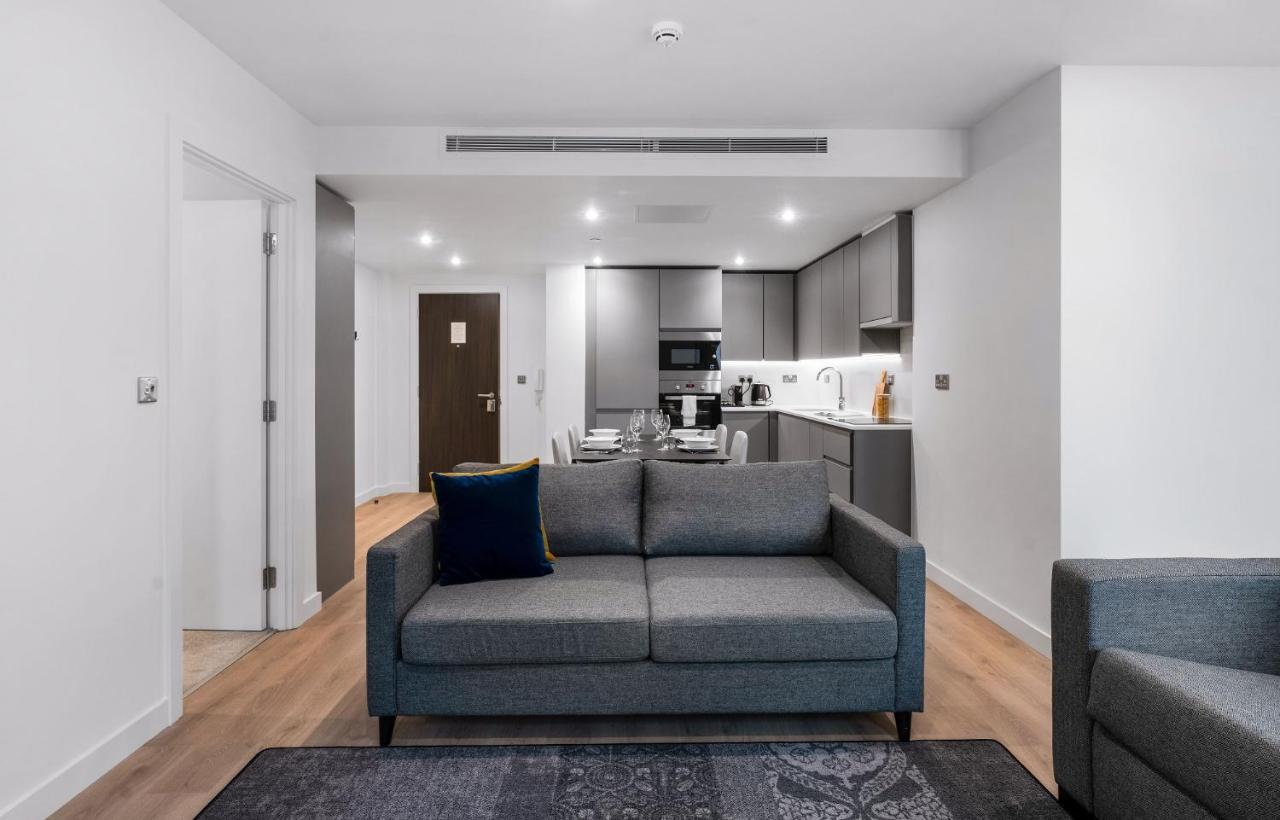 Citysuites 2 Aparthotel Mánchester Exterior foto The photo shows a modern interior space, likely a living area or apartment. In the foreground, there is a gray sofa with a blue cushion positioned on a textured rug. To the back, there is an open kitchen with sleek cabinetry and appliances, visible a