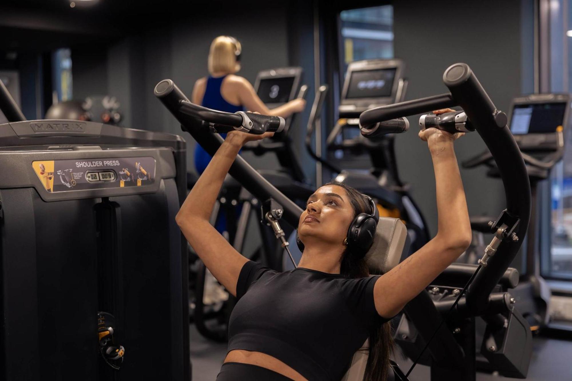 Citysuites 2 Aparthotel Mánchester Exterior foto The photo shows a gym setting with two individuals engaged in physical exercise. In the foreground, a woman is using a chest press machine while wearing headphones, focusing on her workout. She appears to be in a seated position, pushing the handles 